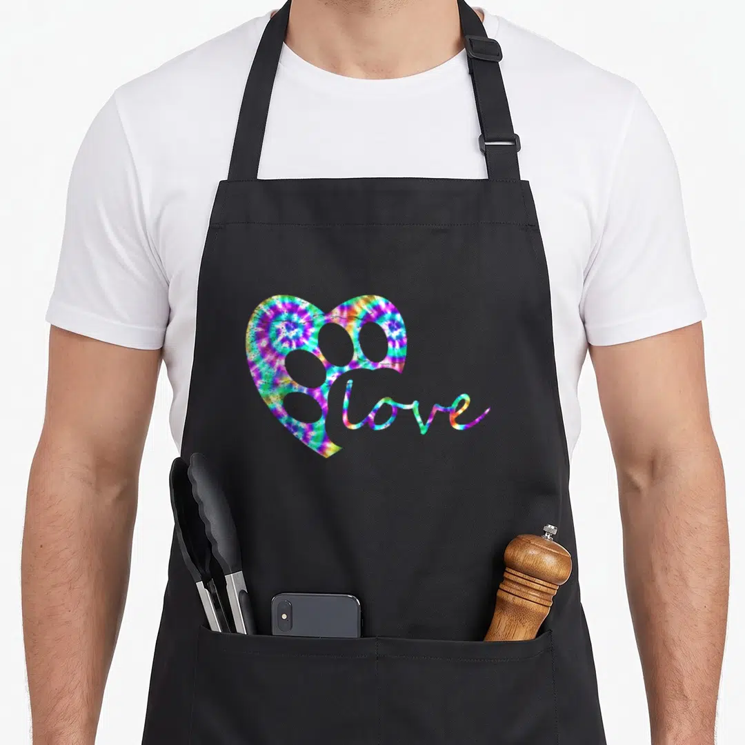 Dog tie-dye apron - Tie-dye heart with paw cutouts and love script in blue purple green yellow pink — shown on a black cotton canvas apron against a white background