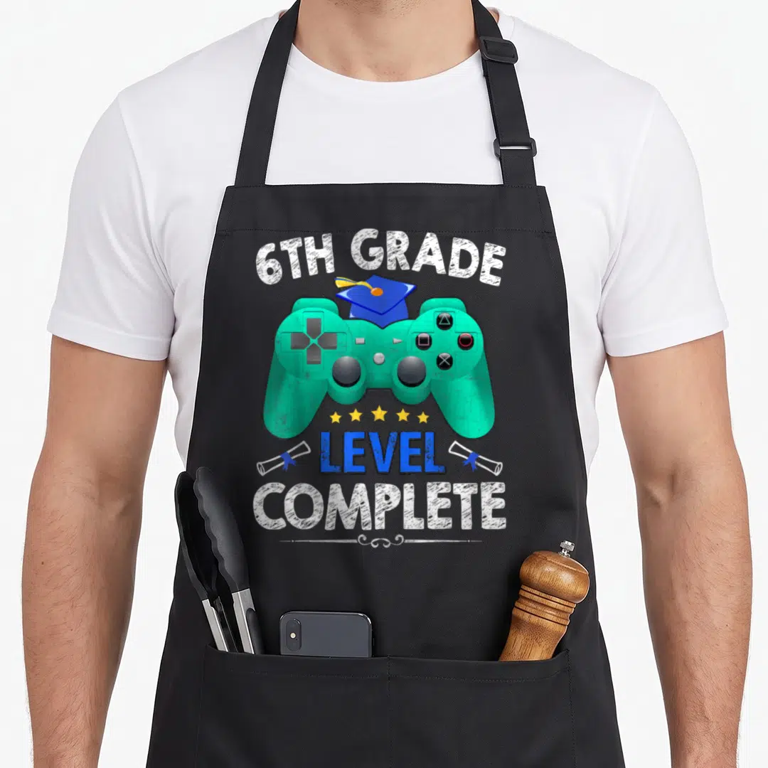 6th grade girls apron - Teal game controller with blue grad cap, 5 stars, and 6TH GRADE LEVEL COMPLETE text — shown on a black cotton canvas apron against a white background