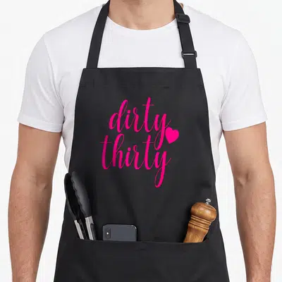 Hot funny dirty aprons - Pink script text reading dirty thirty with a heart icon — shown on a black cotton canvas apron against a white background