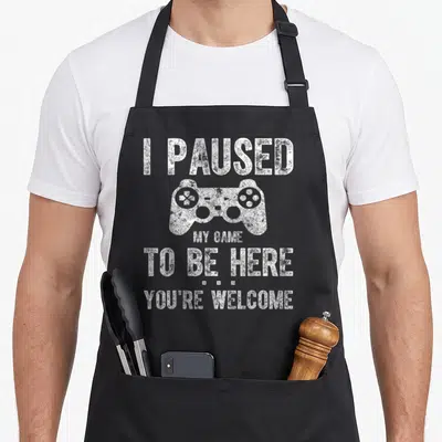 Gamer paused game aprons - White game controller between stacked text reading I PAUSED MY GAME TO BE HERE YOU'RE WELCOME — shown on a black cotton canvas apron against a white background