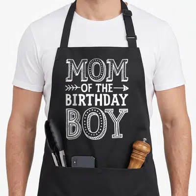 Mama mother's apron - White stacked typography reading MOM OF THE BIRTHDAY BOY with a horizontal arrow on line two — shown on a black cotton canvas apron against a white background