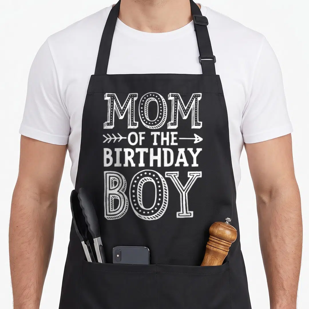 Mama mother's apron - White stacked typography reading MOM OF THE BIRTHDAY BOY with a horizontal arrow on line two — shown on a black cotton canvas apron against a white background