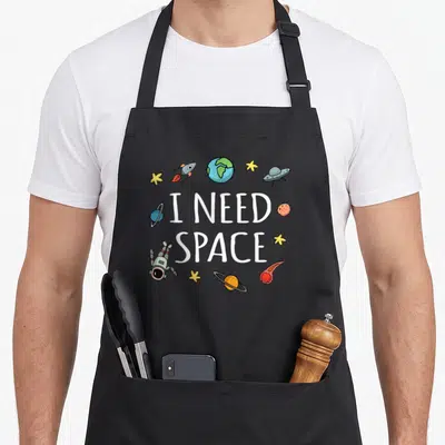Space astronomy science apron - I NEED SPACE text surrounded by cartoon astronaut, planets, stars, rocket, and UFO — shown on a black cotton canvas apron against a white background