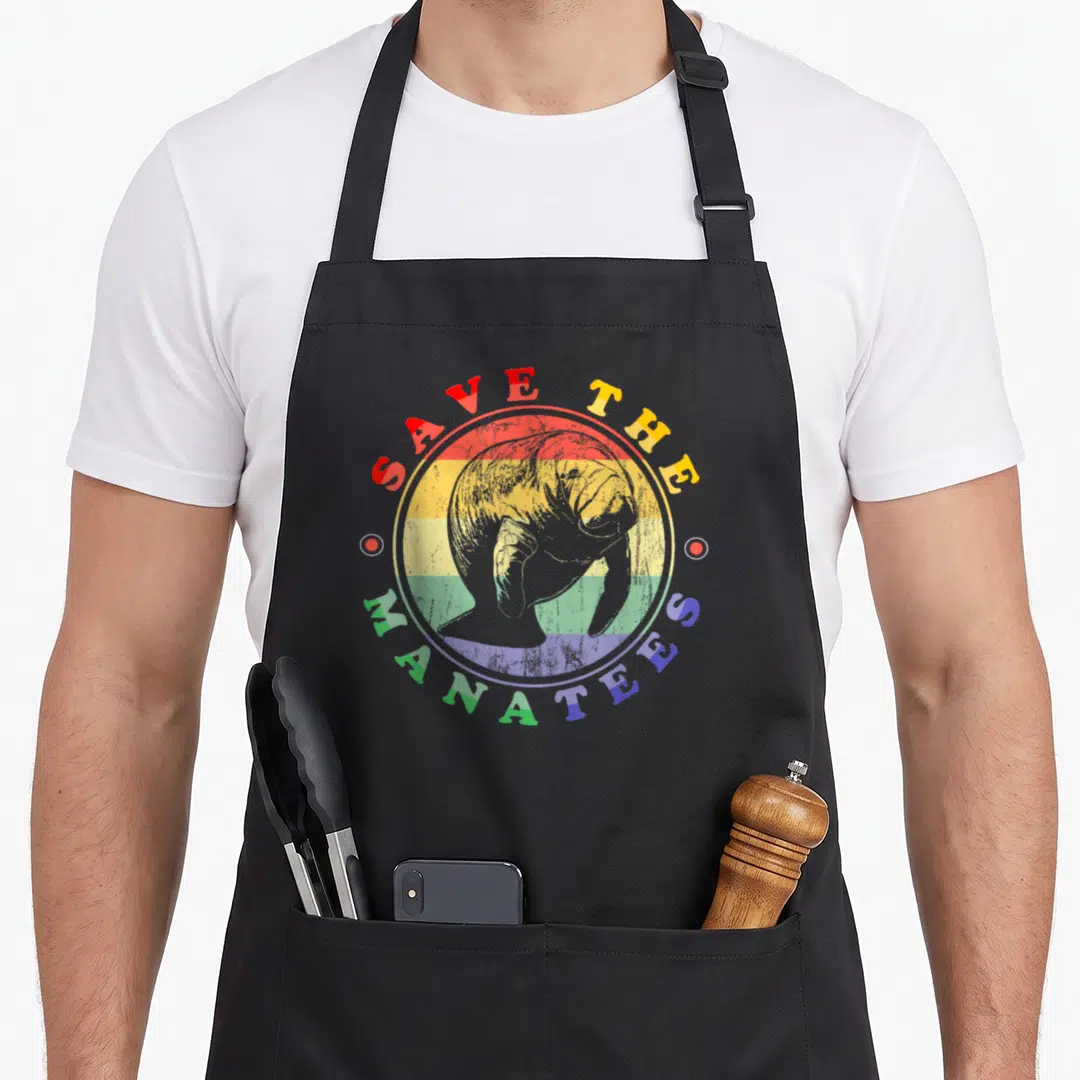 Manatee animal apron - Rainbow striped circle with manatee illustration and arching text — shown on a black cotton canvas apron against a white background