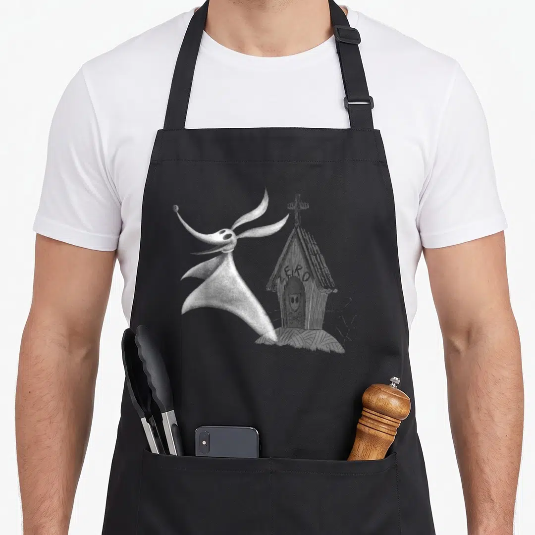 Zero dog apron - White line-art ghost beside a wooden shack with a cross on textured ground — shown on a black cotton canvas apron against a white background