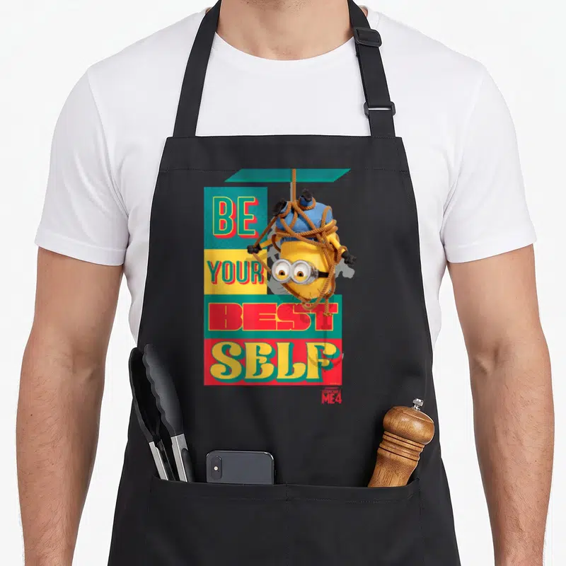 Despicable banana addict apron - Upside down yellow Minion tied in rope over Be Your Best Self text on colored blocks — shown on a black cotton canvas apron against a white background