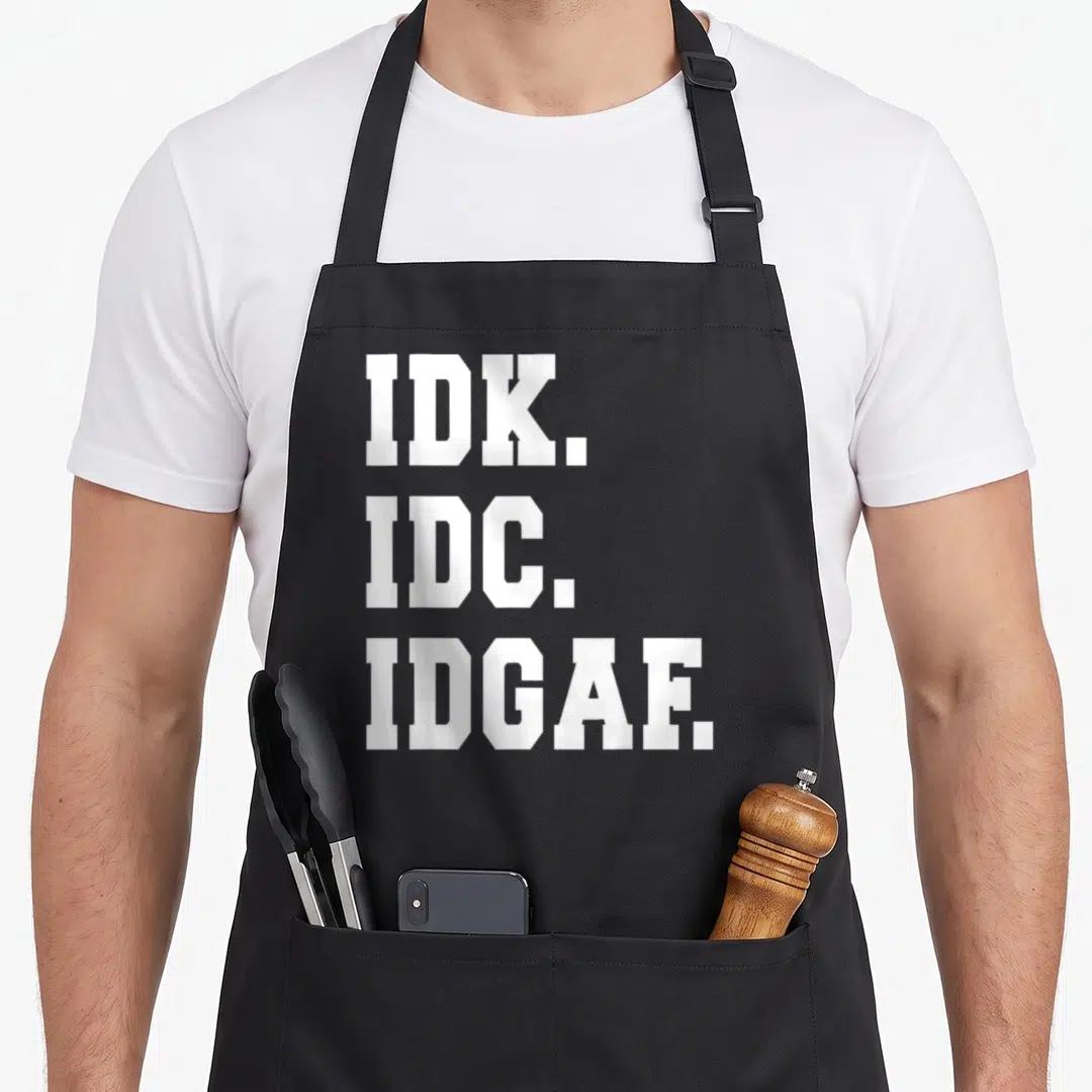 Care idgaf apron - White block letters reading IDK IDC IDGAF stacked vertically on grey background — shown on a black cotton canvas apron against a white background