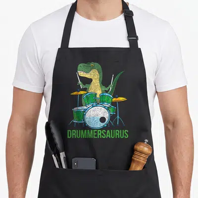 Musician t-rex apron - Green cartoon T-Rex holding drumsticks behind a kit with DRUMMERSAURUS text below — shown on a black cotton canvas apron against a white background