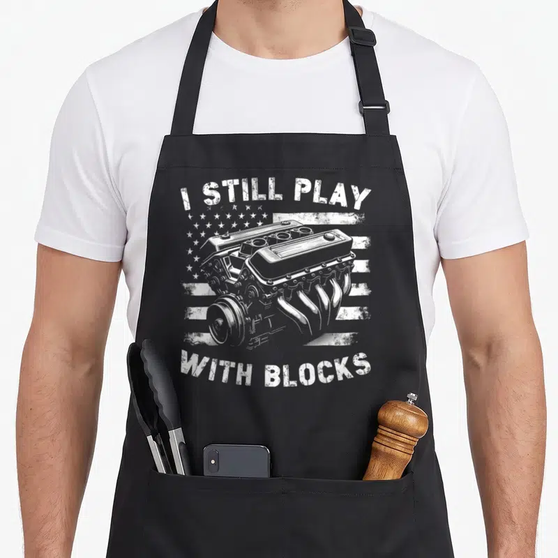 Play garage apron - White distressed engine illustration with arched text I Still Play With Blocks over a stylized flag — shown on a black cotton canvas apron against a white background