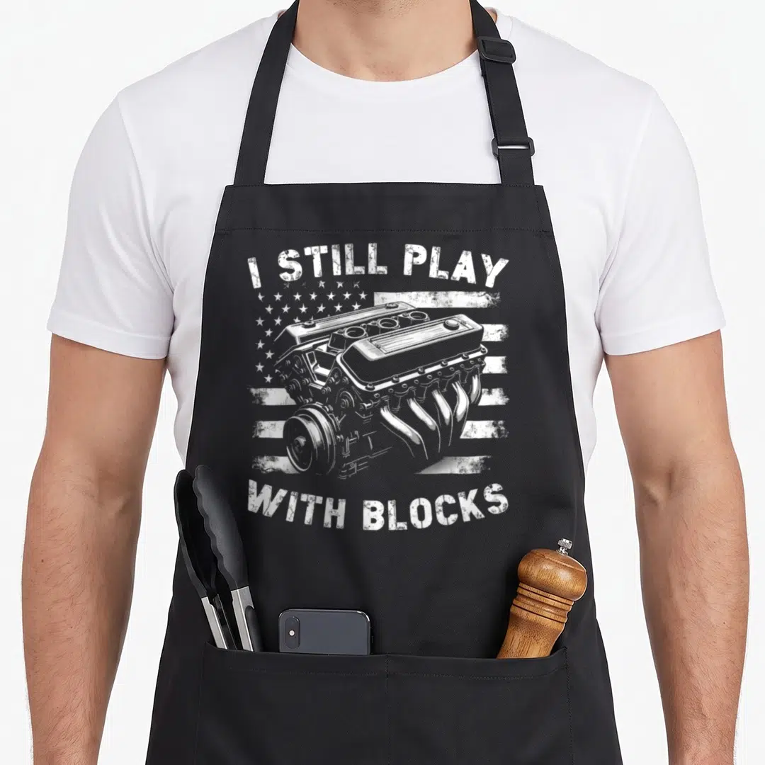Play garage apron - White distressed engine illustration with arched text I Still Play With Blocks over a stylized flag — shown on a black cotton canvas apron against a white background