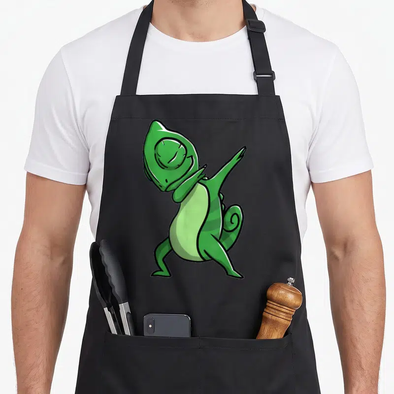 Dabbing chameleon apron - Green cartoon chameleon in a dance pose with light green belly and striped tail — shown on a black cotton canvas apron against a white background