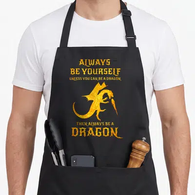 Yourself dragon apron - Cartoon dragon breathing fire with orange and yellow stacked text quote — shown on a black cotton canvas apron against a white background