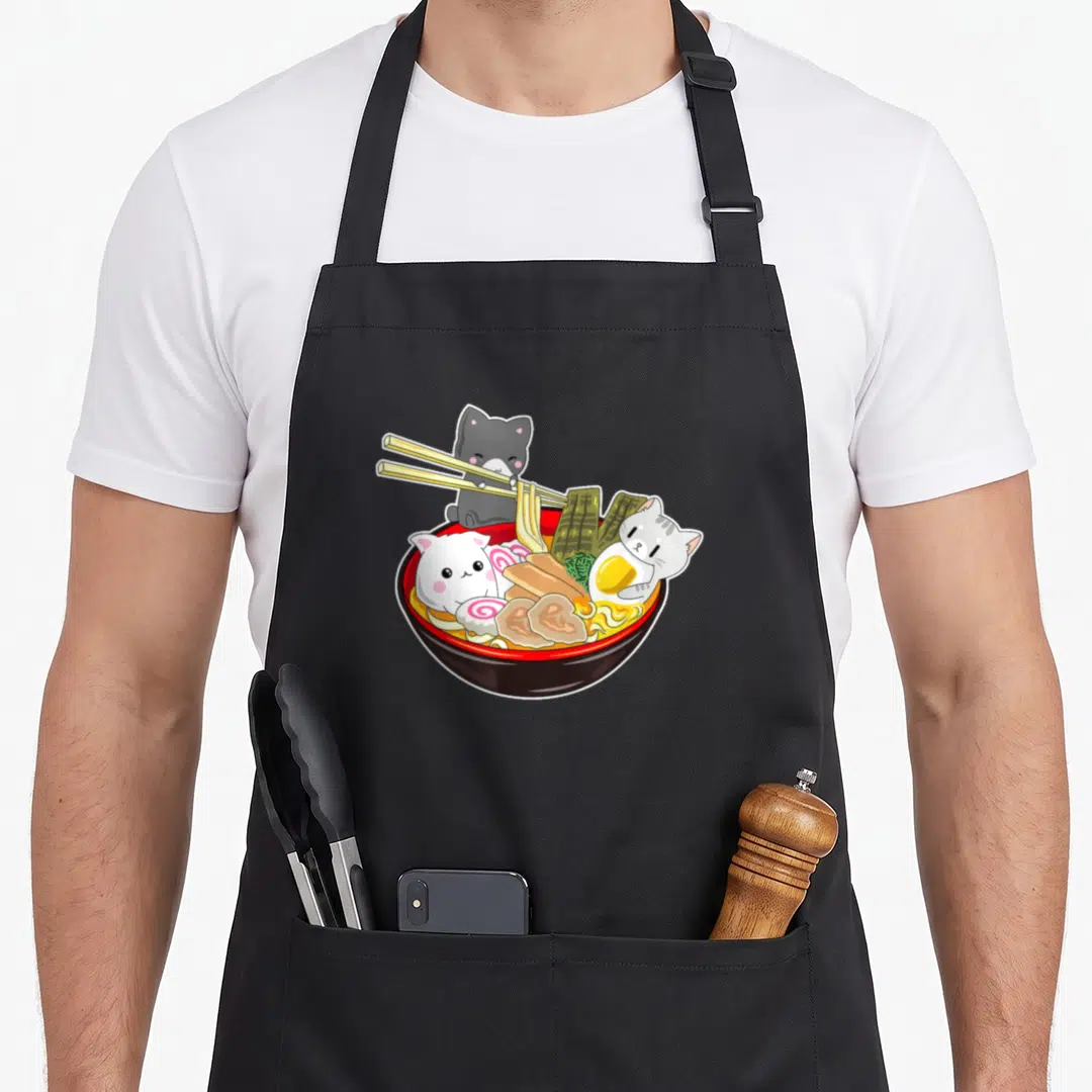Kawaii japanese cat apron - Ramen bowl with three cartoon cats, one holding chopsticks, red and yellow colors — shown on a black cotton canvas apron against a white background