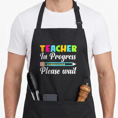 Future progress teacher apron - Colorful TEACHER text above blue pencil with ten small apples and white cursive — shown on a black cotton canvas apron against a white background