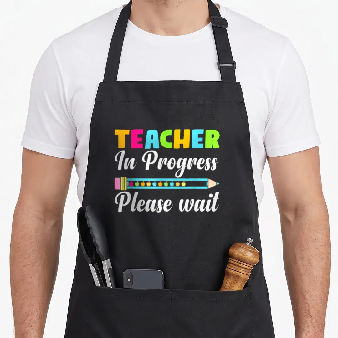 Future progress teacher apron - Colorful TEACHER text above blue pencil with ten small apples and white cursive — shown on a black cotton canvas apron against a white background