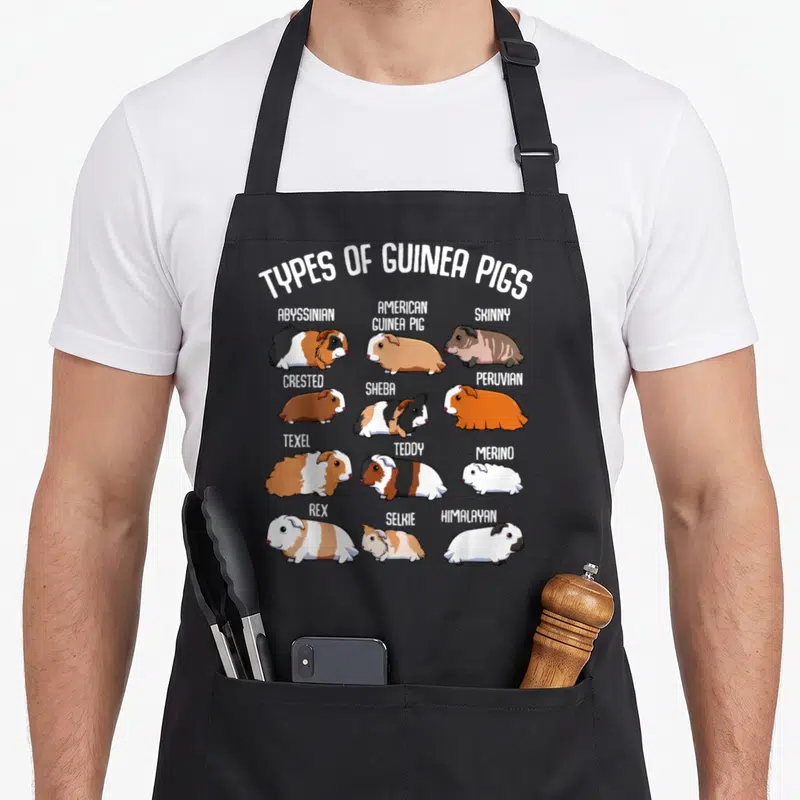 Guinea household aprons - Twelve cartoon guinea pigs labeled with breed names like Abyssinian, Texel, and Himalayan — shown on a black cotton canvas apron against a white background