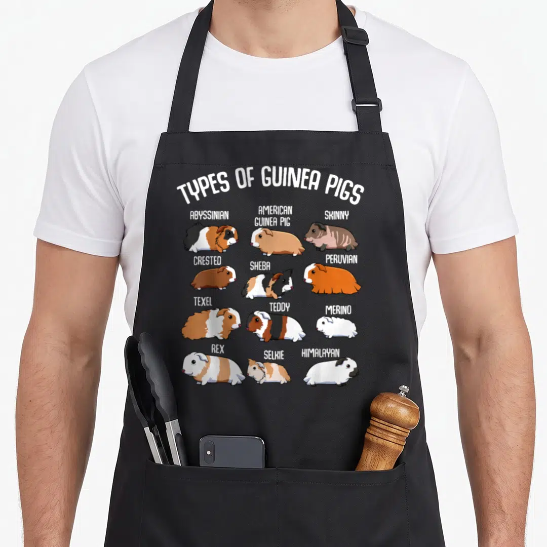Guinea household aprons - Twelve cartoon guinea pigs labeled with breed names like Abyssinian, Texel, and Himalayan — shown on a black cotton canvas apron against a white background
