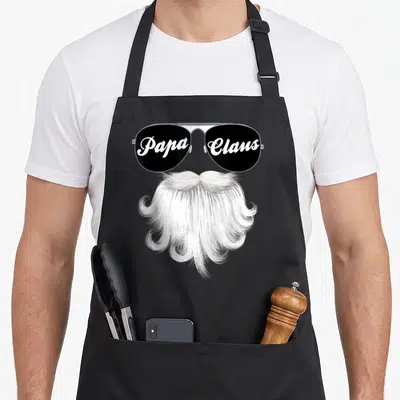 Claus grandad aprons - Papa Claus sunglasses text over white beard in black and white realistic style — shown on a black cotton canvas apron against a white background
