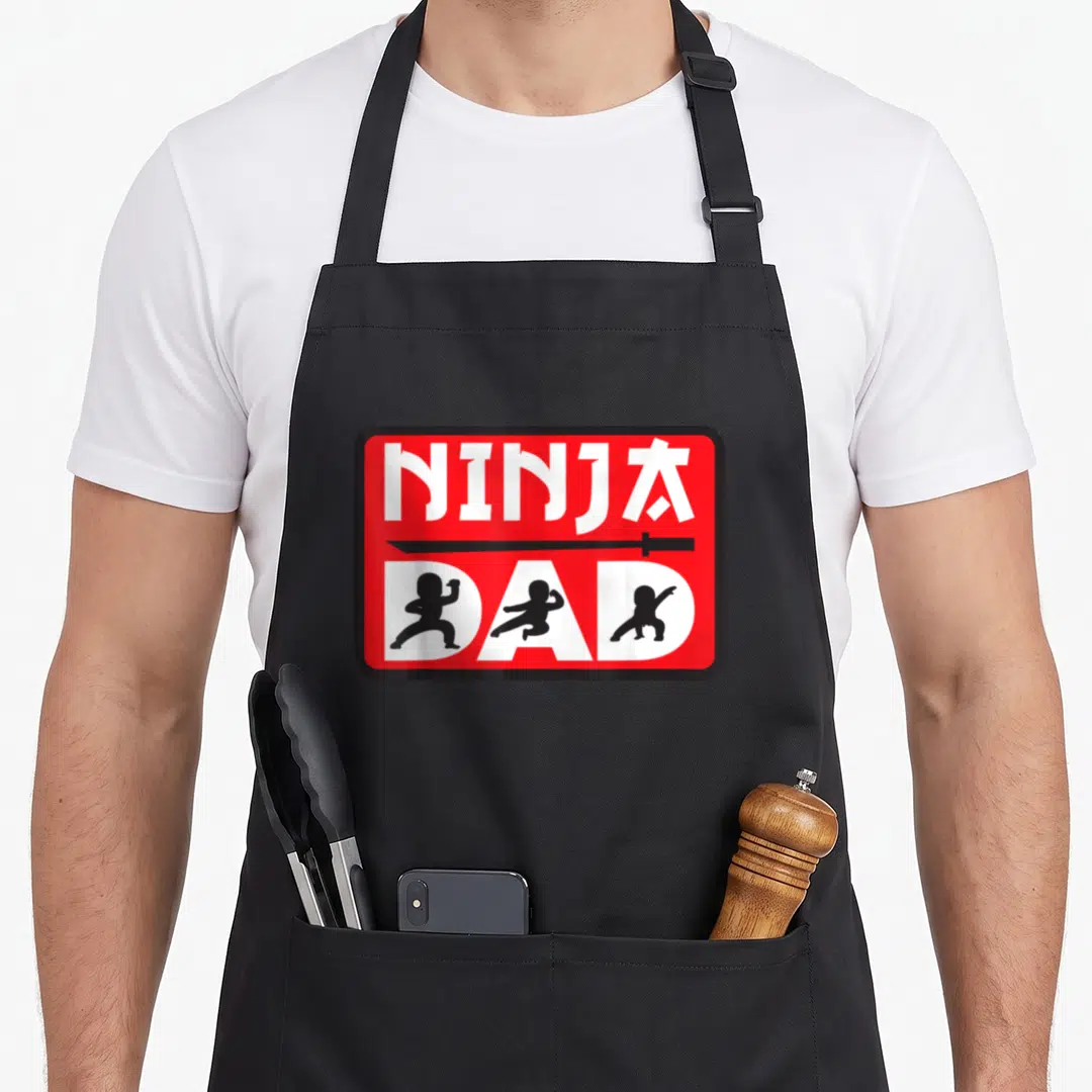 Ninja family aprons - Red badge with NINJA DAD text, black sword, and three ninja silhouettes inside white letters — shown on a black cotton canvas apron against a white background