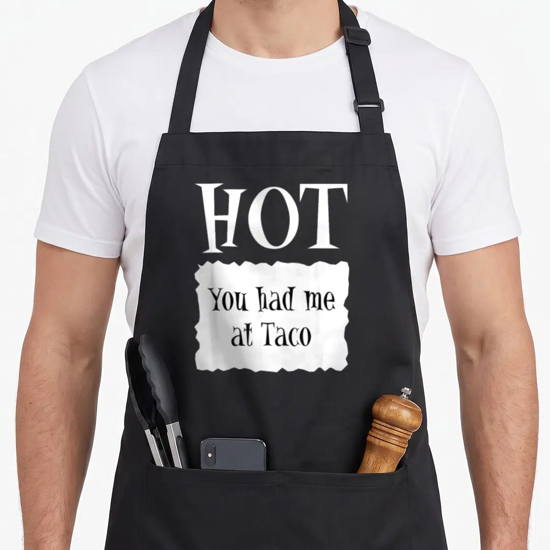 Packet halloween apron - White text HOT above You had me at Taco in black inside torn white paper shape on black — shown on a black cotton canvas apron against a white background