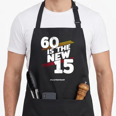 Funny leap 60th birthday apron - White 60 is the new 15 text with yellow orange and red speed lines and hashtag — shown on a black cotton canvas apron against a white background