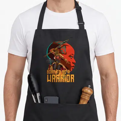 Okoye novelty apron - Woman warrior holding spear in front of red profile face with Born To Be A Warrior text — shown on a black cotton canvas apron against a white background