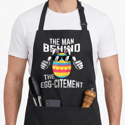 Behind easter egg apron - Cartoon Easter egg with sunglasses, bunny ears, and thumbs up flanked by white text — shown on a black cotton canvas apron against a white background