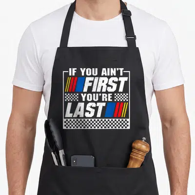 Motor car apron - Racing text IF YOU AIN'T FIRST YOU'RE LAST with stripes and checkered patterns — shown on a black cotton canvas apron against a white background