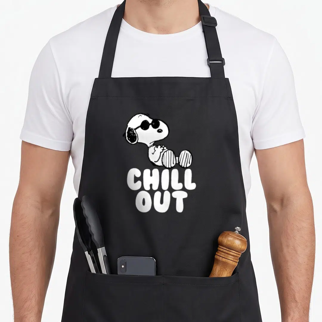 Chill crew apron - Snoopy wearing sunglasses sitting above white CHILL OUT text in black and white cartoon style — shown on a black cotton canvas apron against a white background