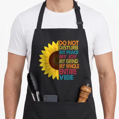 Disturb peace joy grind apron - Half sunflower next to stacked DO NOT DISTURB MY PEACE MY JOY MY GRIND MY WHOLE ENTIRE — shown on a black cotton canvas apron against a white background