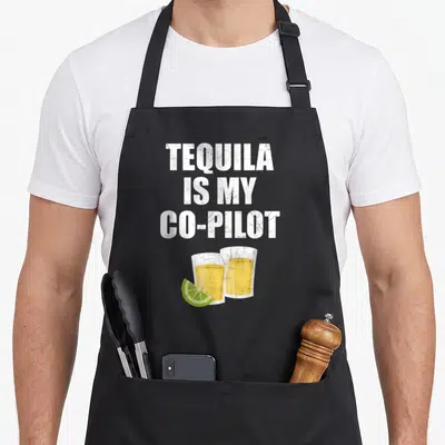 Tequila chef and co aprons - Distressed text TEQUILA IS MY CO-PILOT above two yellow shot glasses and green lime wedge — shown on a black cotton canvas apron against a white background