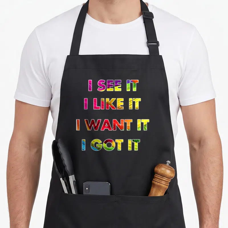 See i love watching apron - Stacked I See It I Like It I Want It I Got It text in pink yellow green red and blue — shown on a black cotton canvas apron against a white background