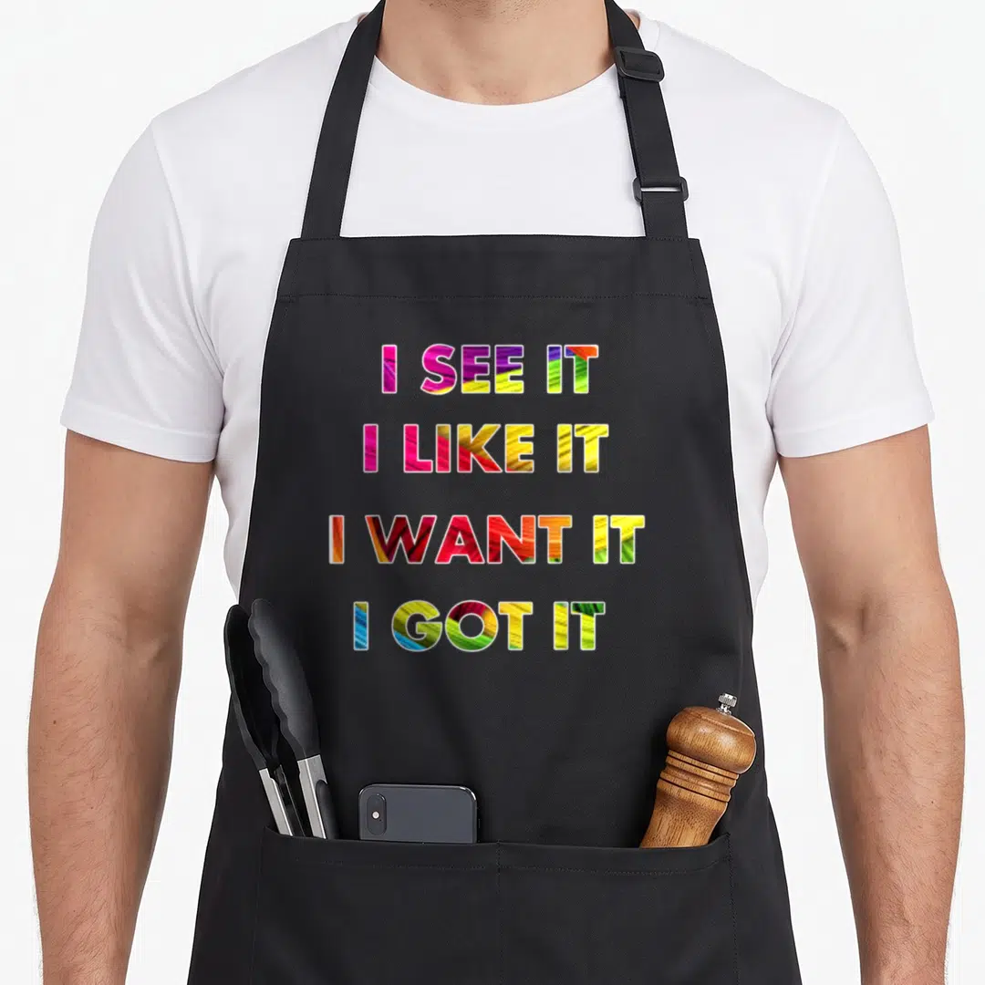 See i love watching apron - Stacked I See It I Like It I Want It I Got It text in pink yellow green red and blue — shown on a black cotton canvas apron against a white background