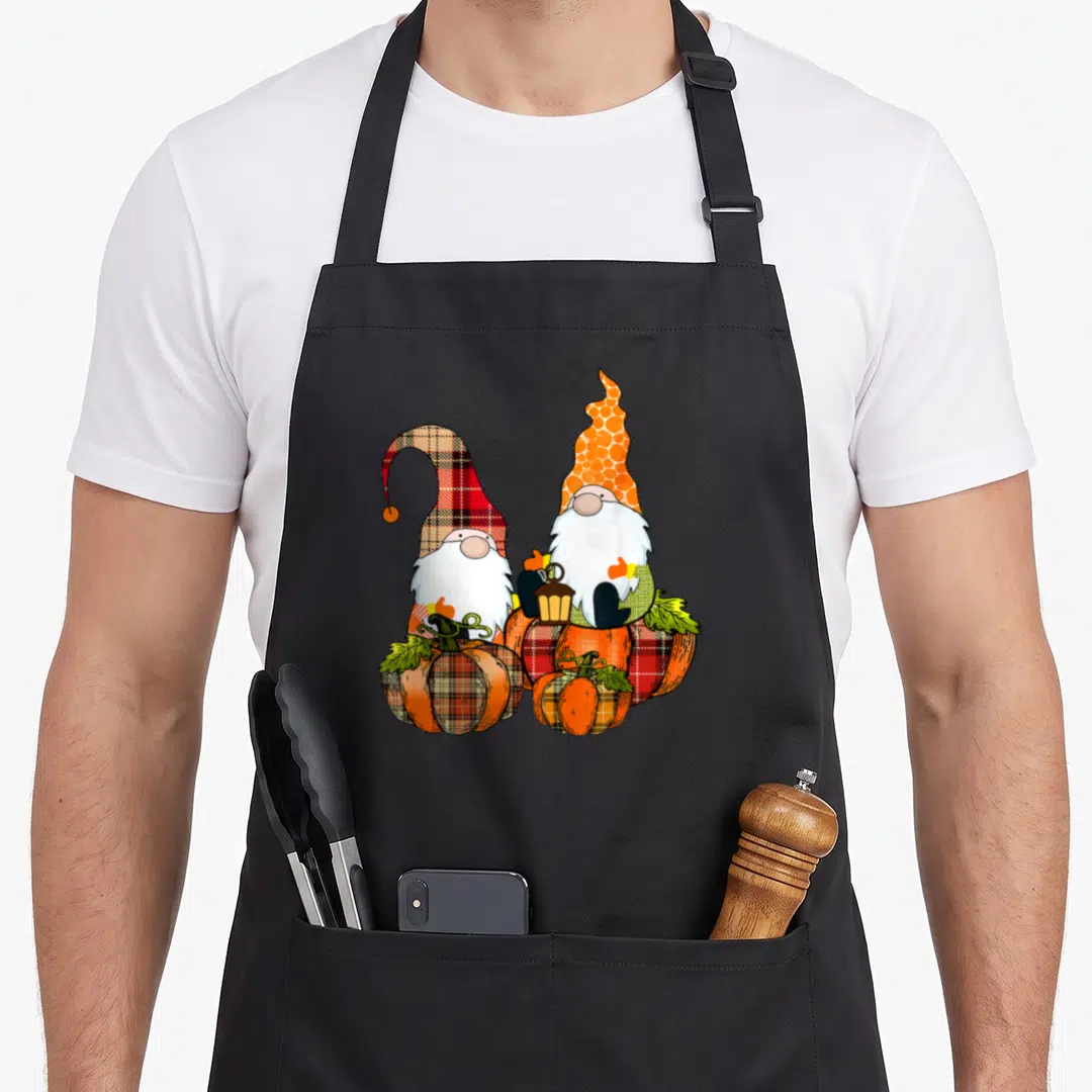 Gnome fall aprons - Two cartoon gnomes sitting behind four plaid pumpkins in orange and red — shown on a black cotton canvas apron against a white background
