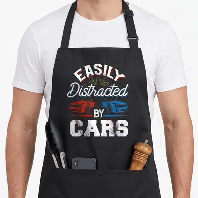 Easily distracted car apron - Arched EASILY Distracted BY CARS text above red and blue facing cars with yellow sunburst — shown on a black cotton canvas apron against a white background