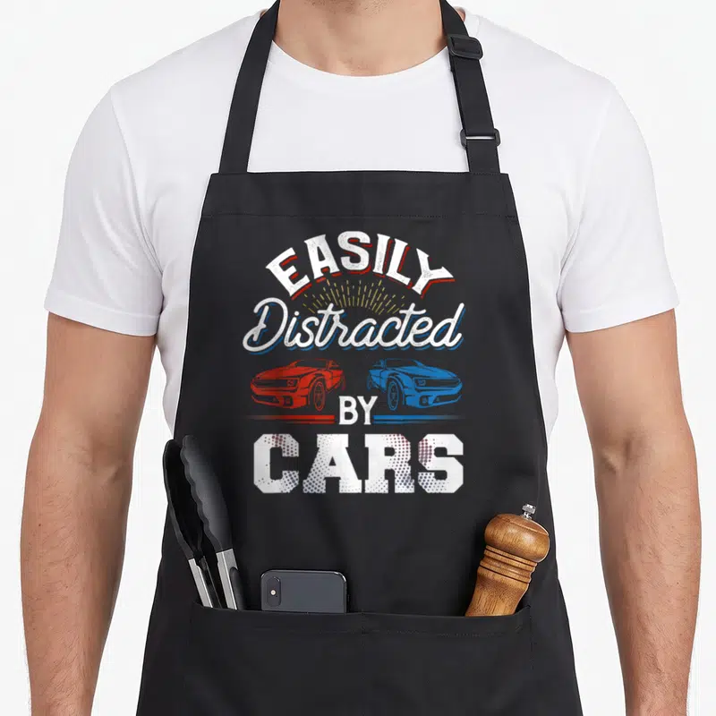 Easily distracted car apron - Arched EASILY Distracted BY CARS text above red and blue facing cars with yellow sunburst — shown on a black cotton canvas apron against a white background