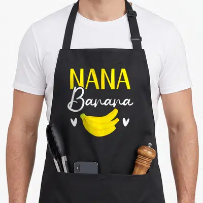 Banana grandma apron - Nana Banana text above yellow bananas with two white hearts — shown on a black cotton canvas apron against a white background