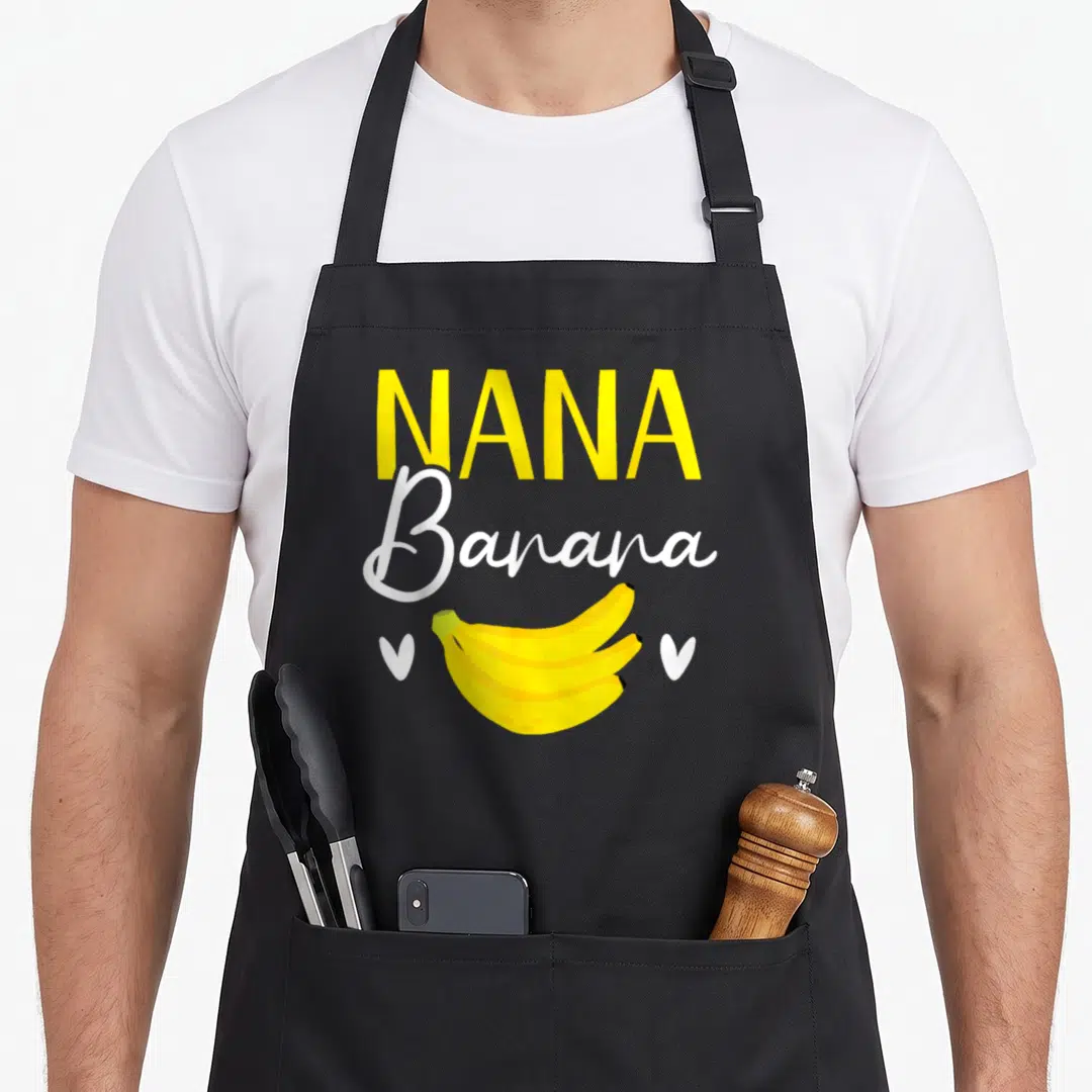 Banana grandma apron - Nana Banana text above yellow bananas with two white hearts — shown on a black cotton canvas apron against a white background