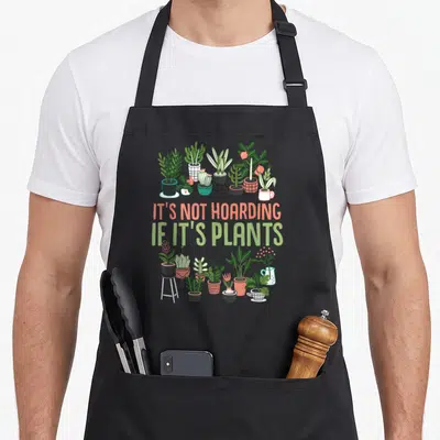 Botanist plant apron - Green and coral potted plants framing text It's Not Hoarding If It's Plants — shown on a black cotton canvas apron against a white background