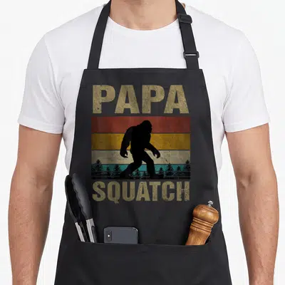Papa yeti apron - Retro bigfoot silhouette over striped sunset with PAPA SQUATCH text in distressed font — shown on a black cotton canvas apron against a white background