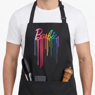 Rainbow drip apron - Barbie script logo with rainbow paint dripping down in pink, orange, yellow, green, blue — shown on a black cotton canvas apron against a white background