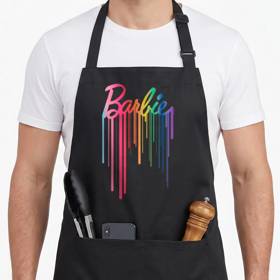 Rainbow drip apron - Barbie script logo with rainbow paint dripping down in pink, orange, yellow, green, blue — shown on a black cotton canvas apron against a white background