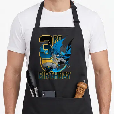 3rd action birthday apron - Leaping Batman character in front of 3RD BIRTHDAY text in blue and yellow — shown on a black cotton canvas apron against a white background