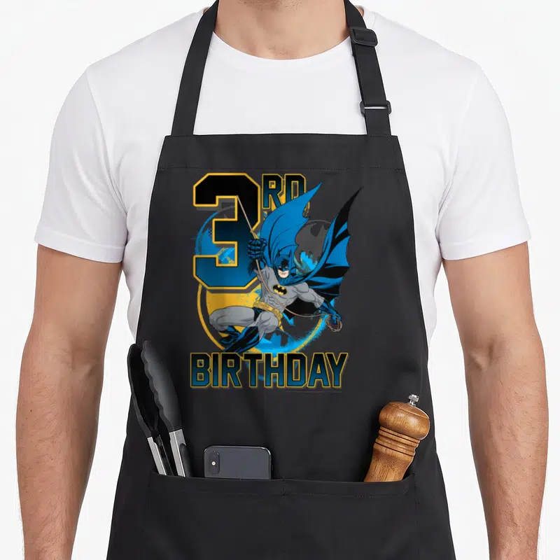3rd action birthday apron - Leaping Batman character in front of 3RD BIRTHDAY text in blue and yellow — shown on a black cotton canvas apron against a white background