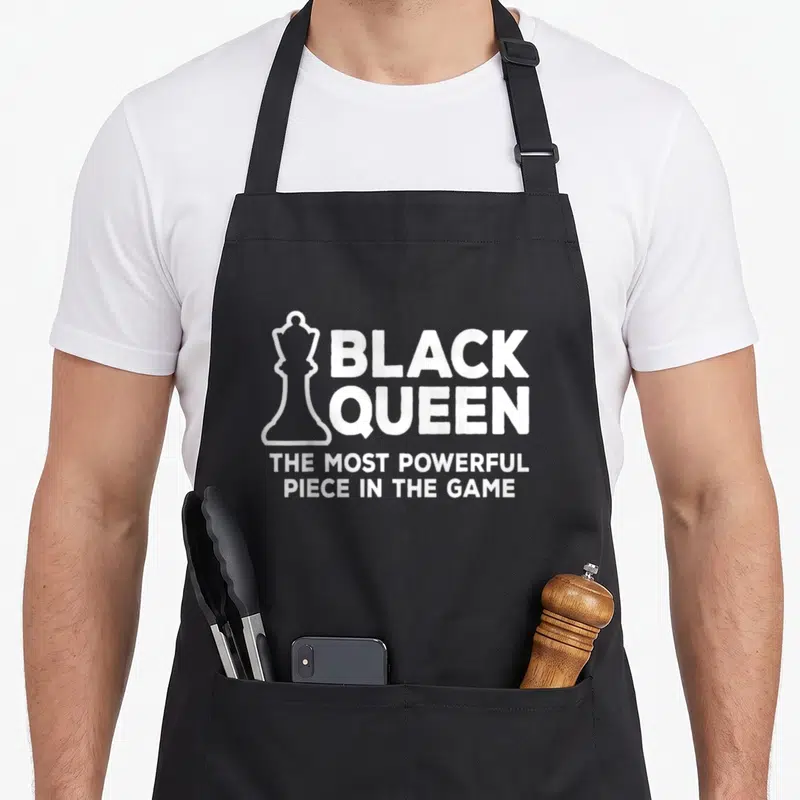 Powerful african american aprons - White chess queen outline next to BLACK QUEEN THE MOST POWERFUL PIECE IN THE GAME — shown on a black cotton canvas apron against a white background