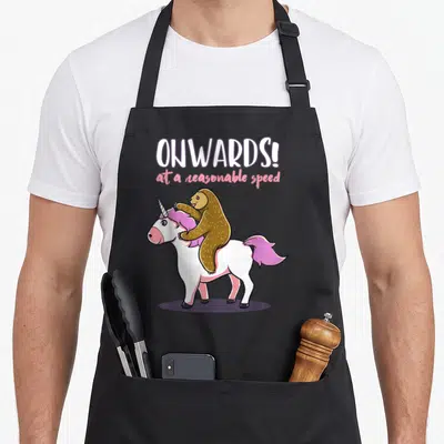 Sloth unicorn apron - ONWARDS! at a reasonable speed text above a brown sloth riding a white unicorn with pink mane — shown on a black cotton canvas apron against a white background
