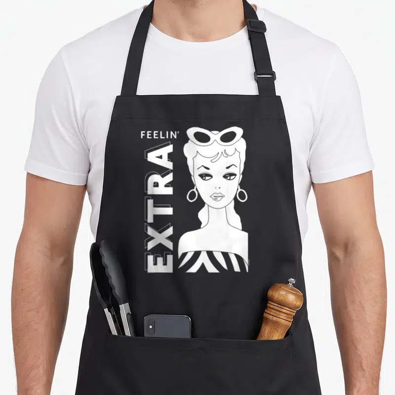 Feelin an extra apron - White line art of a woman with sunglasses and FEELIN EXTRA text — shown on a black cotton canvas apron against a white background