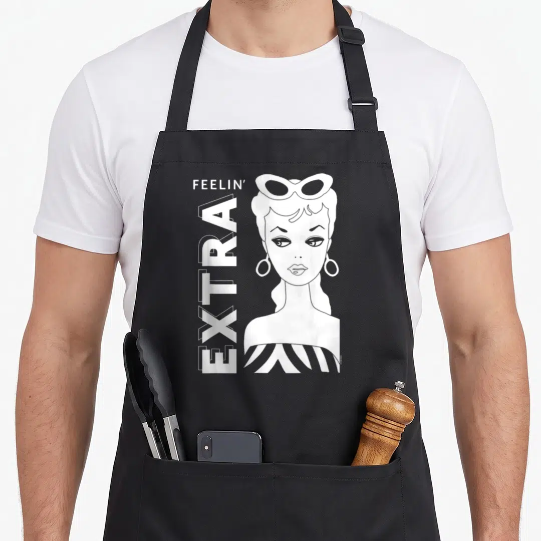 Feelin an extra apron - White line art of a woman with sunglasses and FEELIN EXTRA text — shown on a black cotton canvas apron against a white background