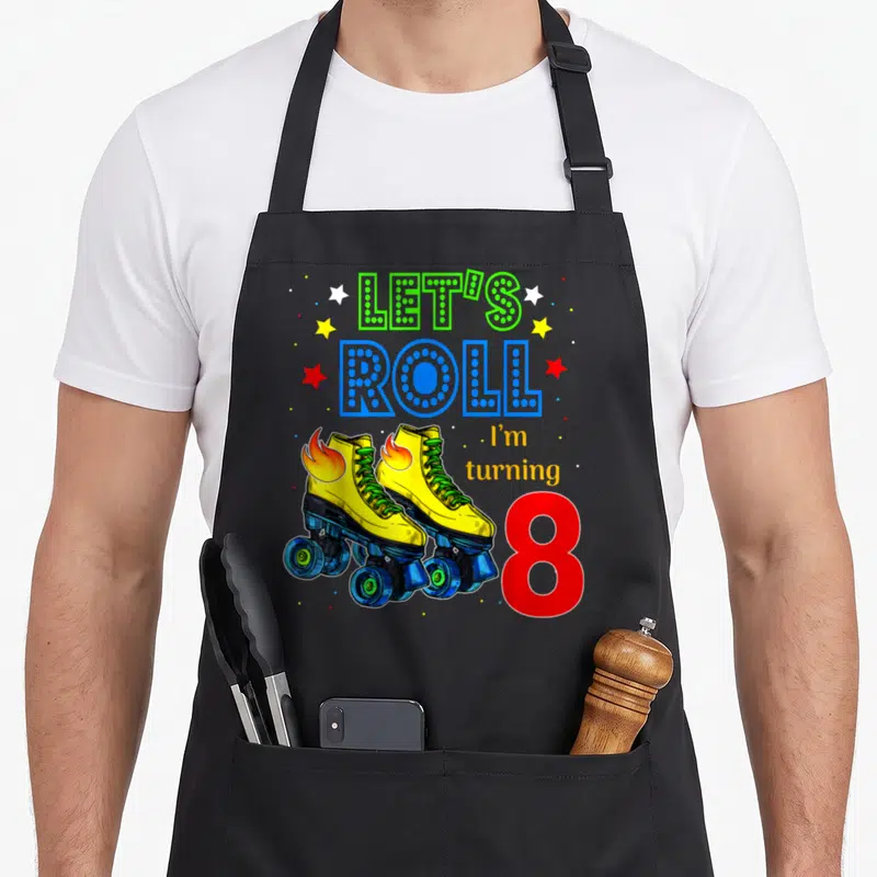Funny roll turning apron - LET'S ROLL text above yellow roller skates with flames and red number 8 — shown on a black cotton canvas apron against a white background