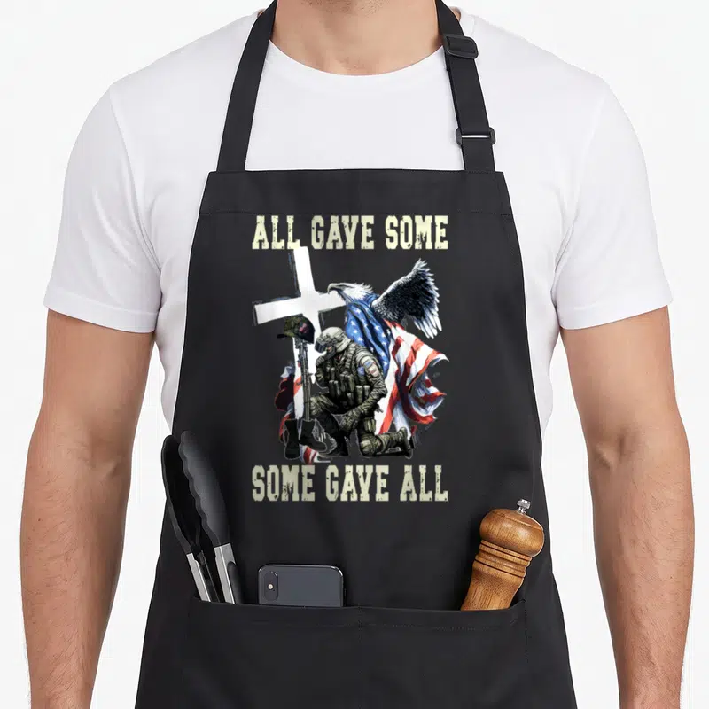 Gave us aprons - Kneeling soldier in olive gear before white cross and US flag with perched eagle and text — shown on a black cotton canvas apron against a white background